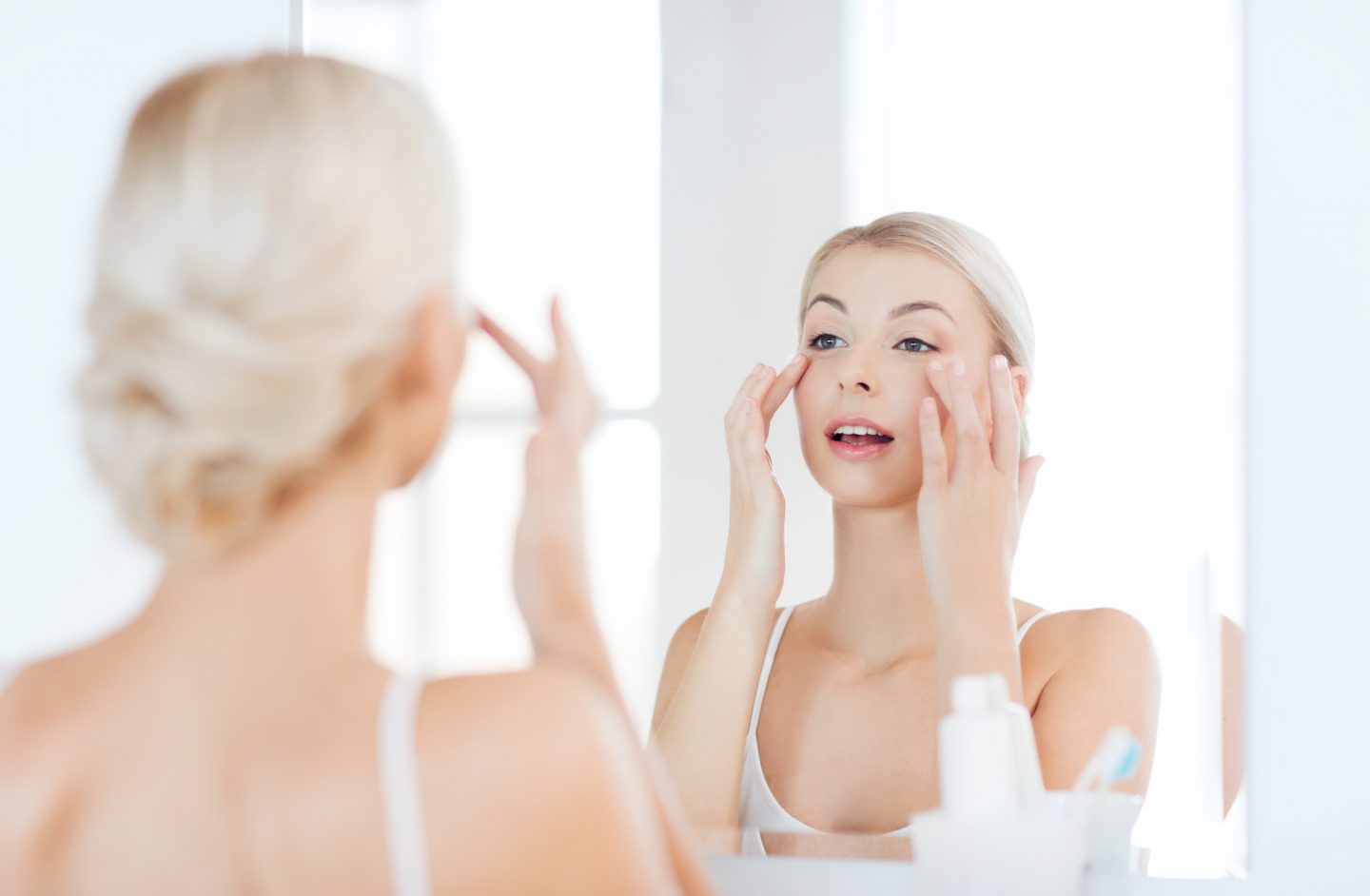Happy blonde woman in front of a mirror applying cream to her face