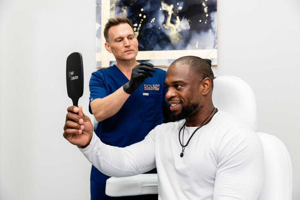 Dr. Beck shows a male sitting in an examination chair where he'll make alterations to his hairline. The patient smiles looking in a hand-held mirror as he gets advice from the best plastic surgeon in Frisco, TX.