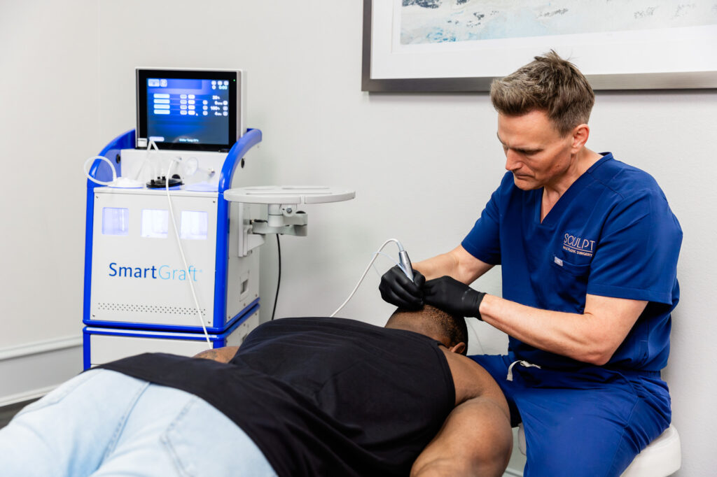 An African American male patient lays face-down on a treatment table as Dr. Beck uses state-of-the-art equipment to perform a FUE hair transplant in Frisco, TX. The procedure is simple, safe, suture-free, and done on an outpatient basis.