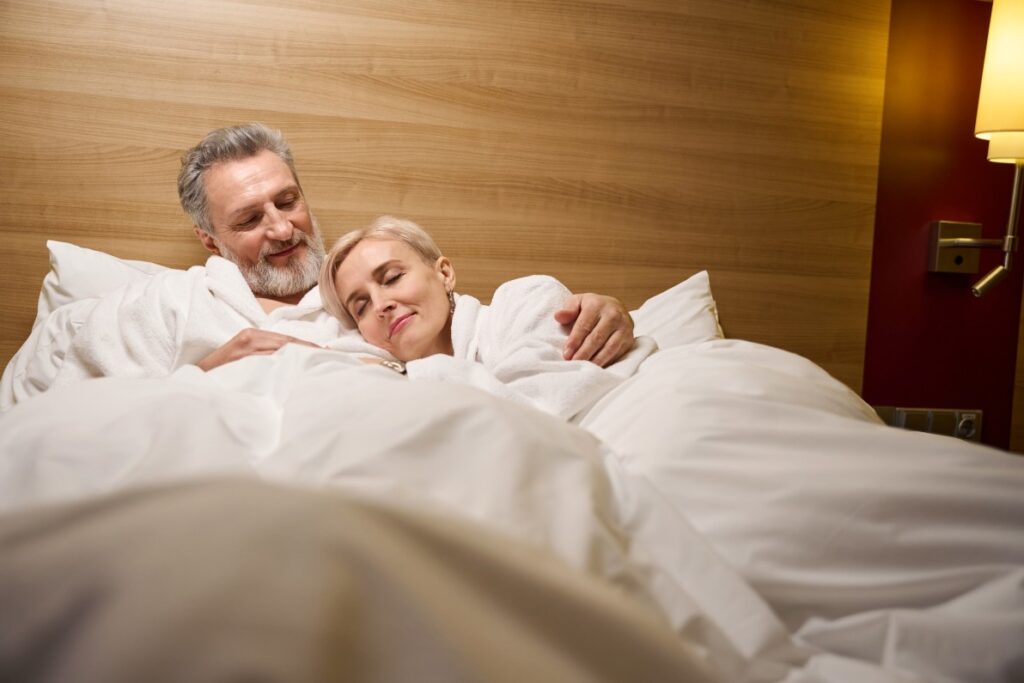 A mature man and woman lie in a cozy bed, snuggling. His PRP injections in Dallas have brought back not only his libido, but his power to satisfy.